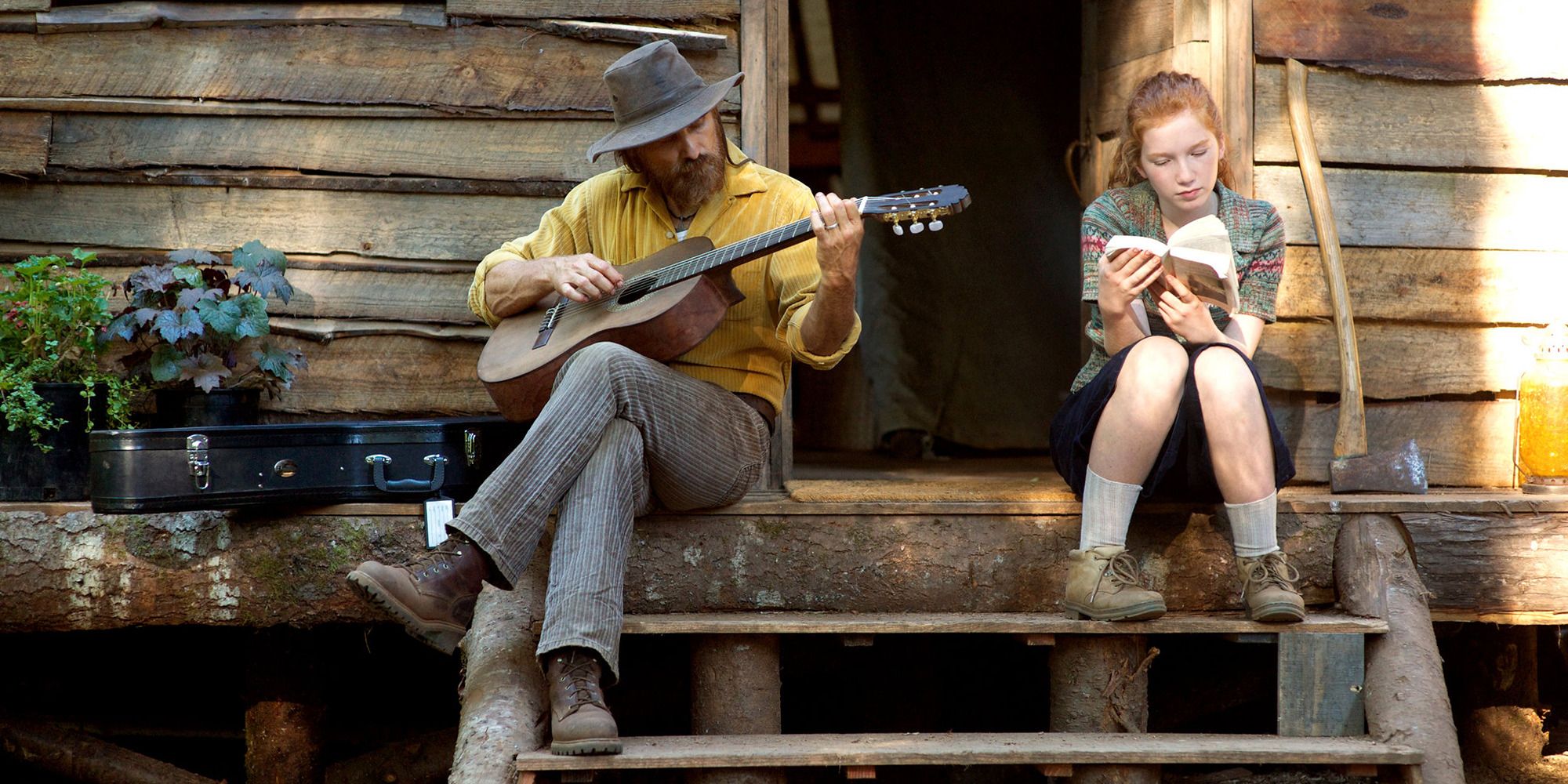 A man playing the guitar on his porch while his daughter reads a book beside him