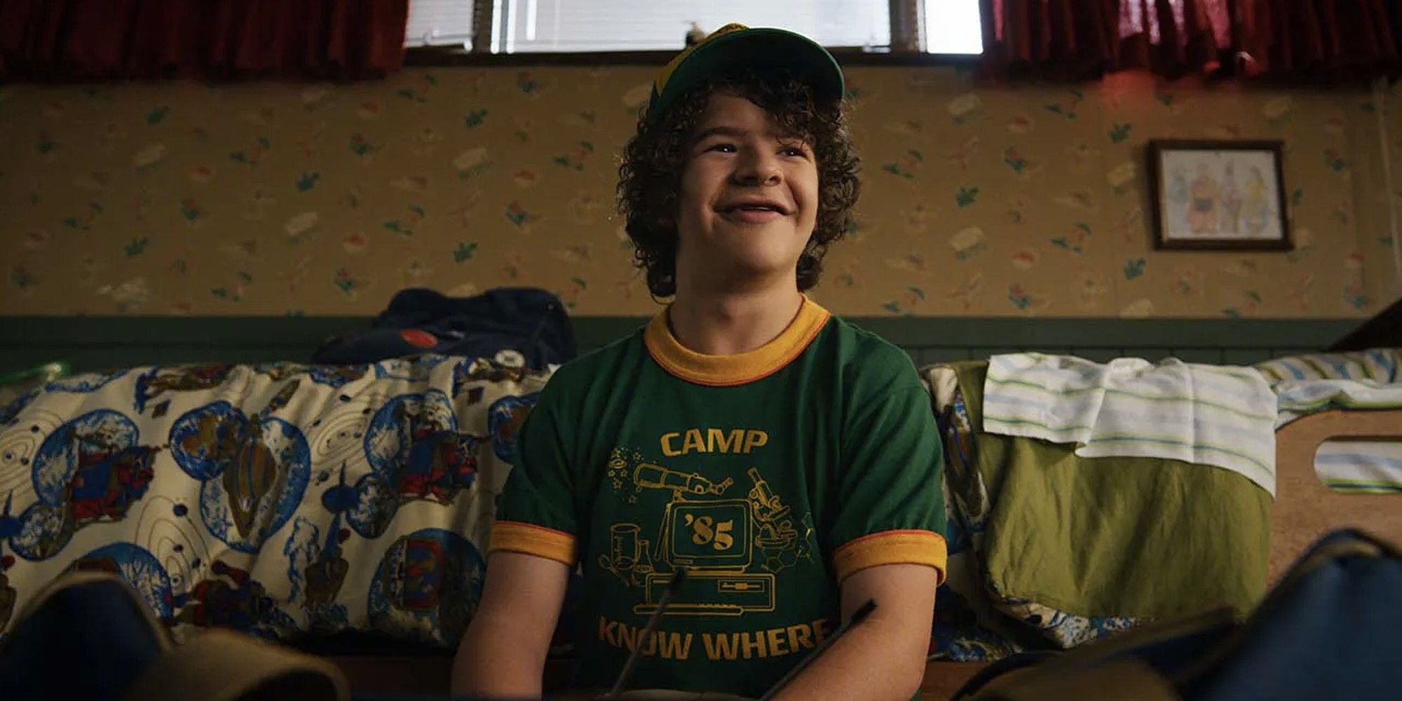 Dustin sits on a couch with a &ldquo;Camp Know Where&rdquo; shirt on and a smile on his face in &lsquo;Stranger Things&rsquo;