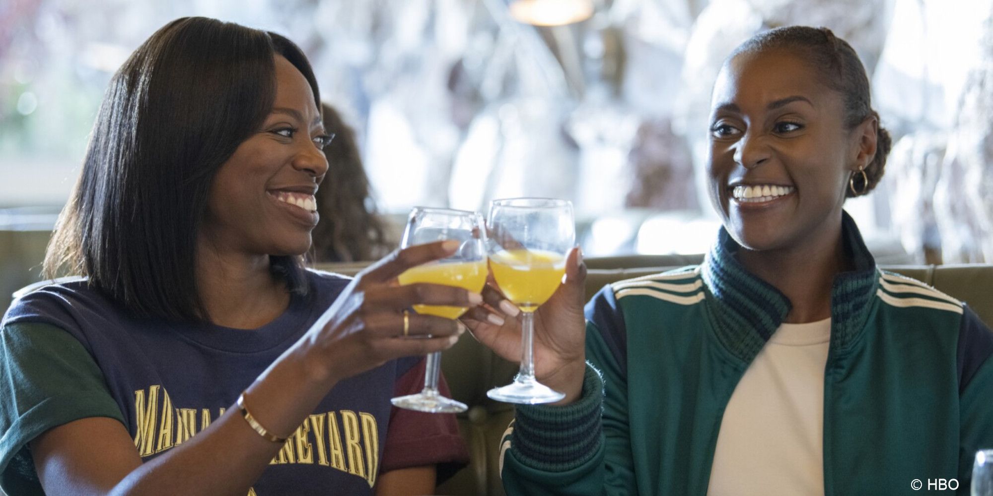 Molly and Issa clink mimosa glasses while smiling at one another in a restaurant booth in Insecure.