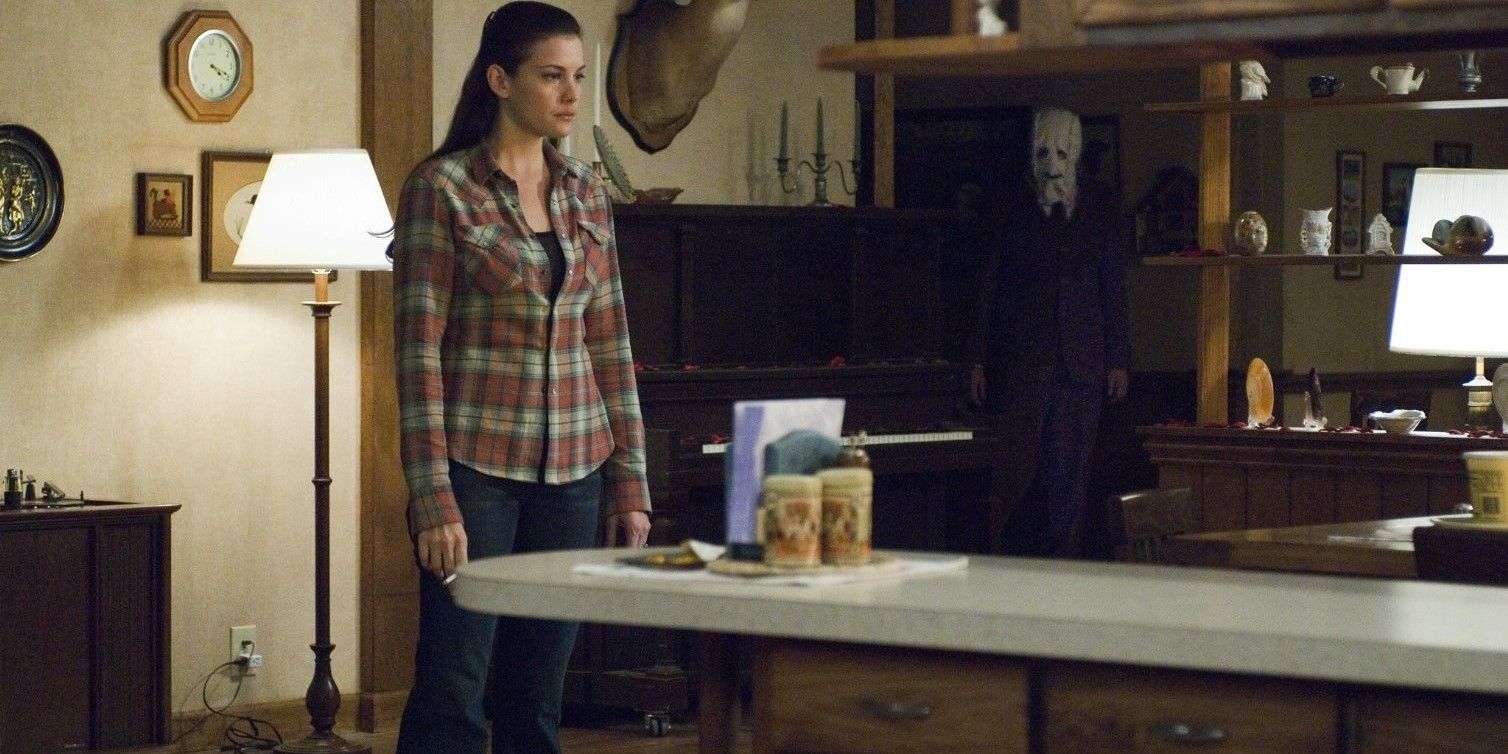 Kristen McKay standing in her home with a masked man lurking in the background in The Strangers