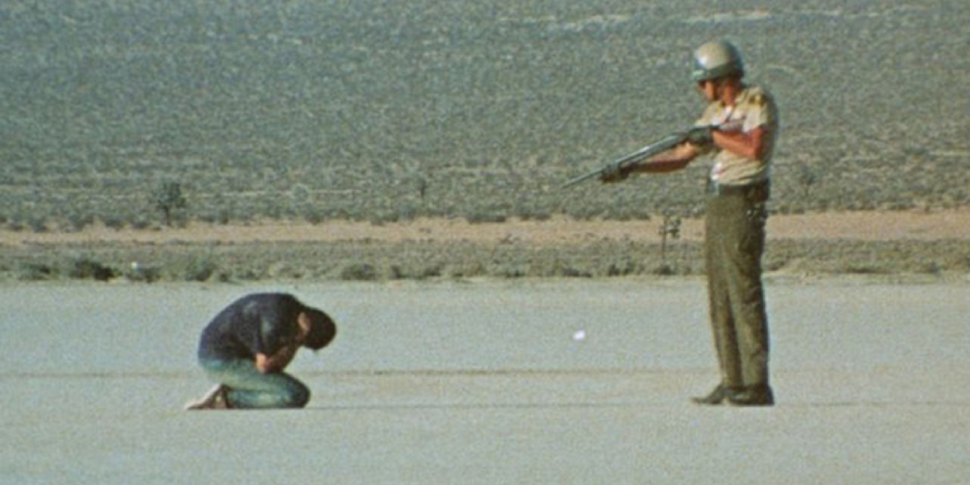 A police officer aims a shotgun at a kneeling man