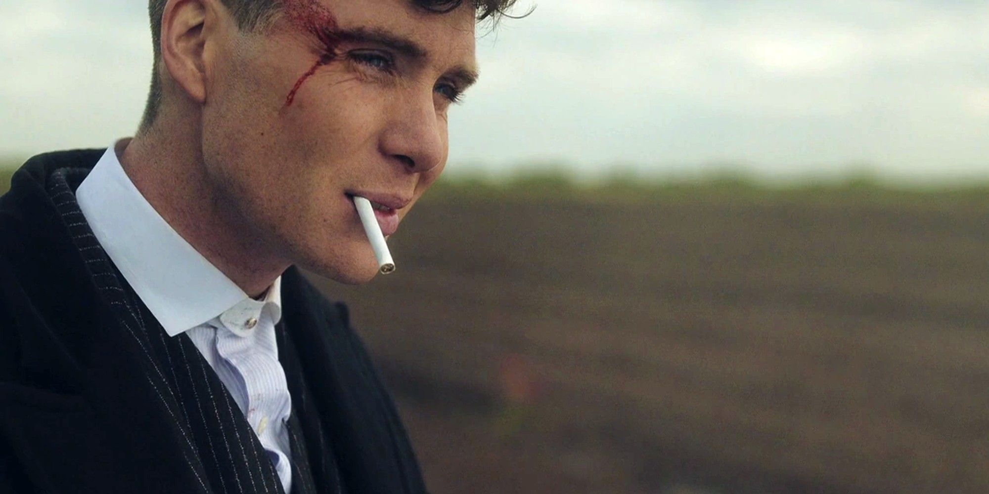 Tommy Shelby smoking and smirking while standing in an open field in Peaky Blinders