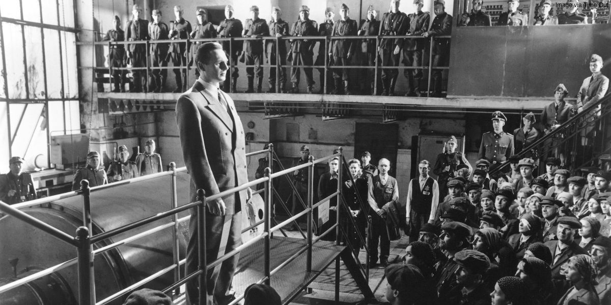 Schindler (Liam Neeson) standing in front of workers and soldiers in a factory