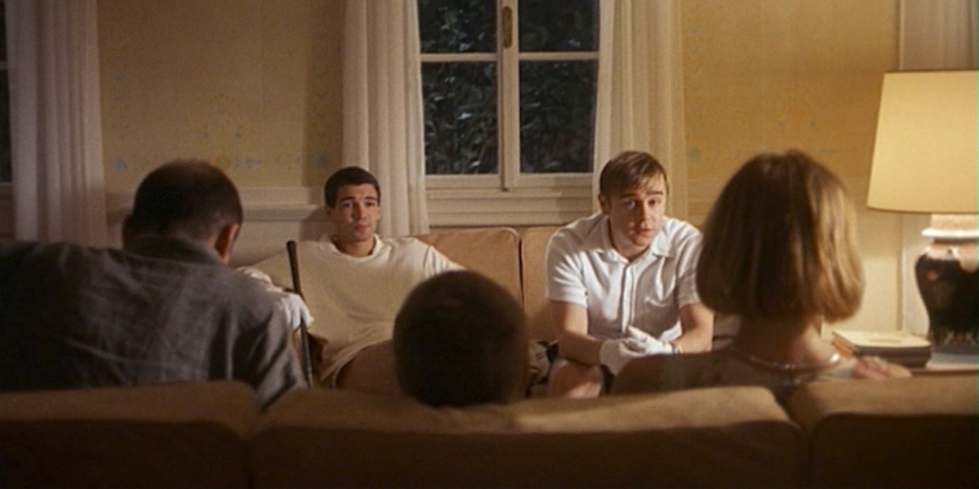 Two young men in white clothes sit on a couch across from a family of three