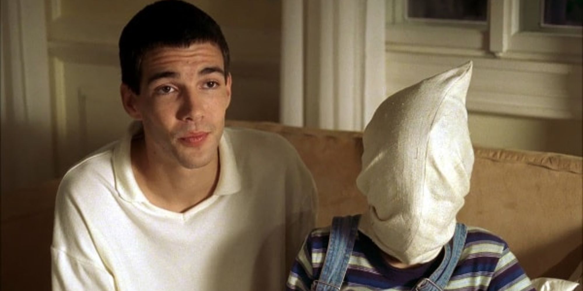 A young man sitting next to a child with a bedsheet cover over the face in Funny Games (1997)
