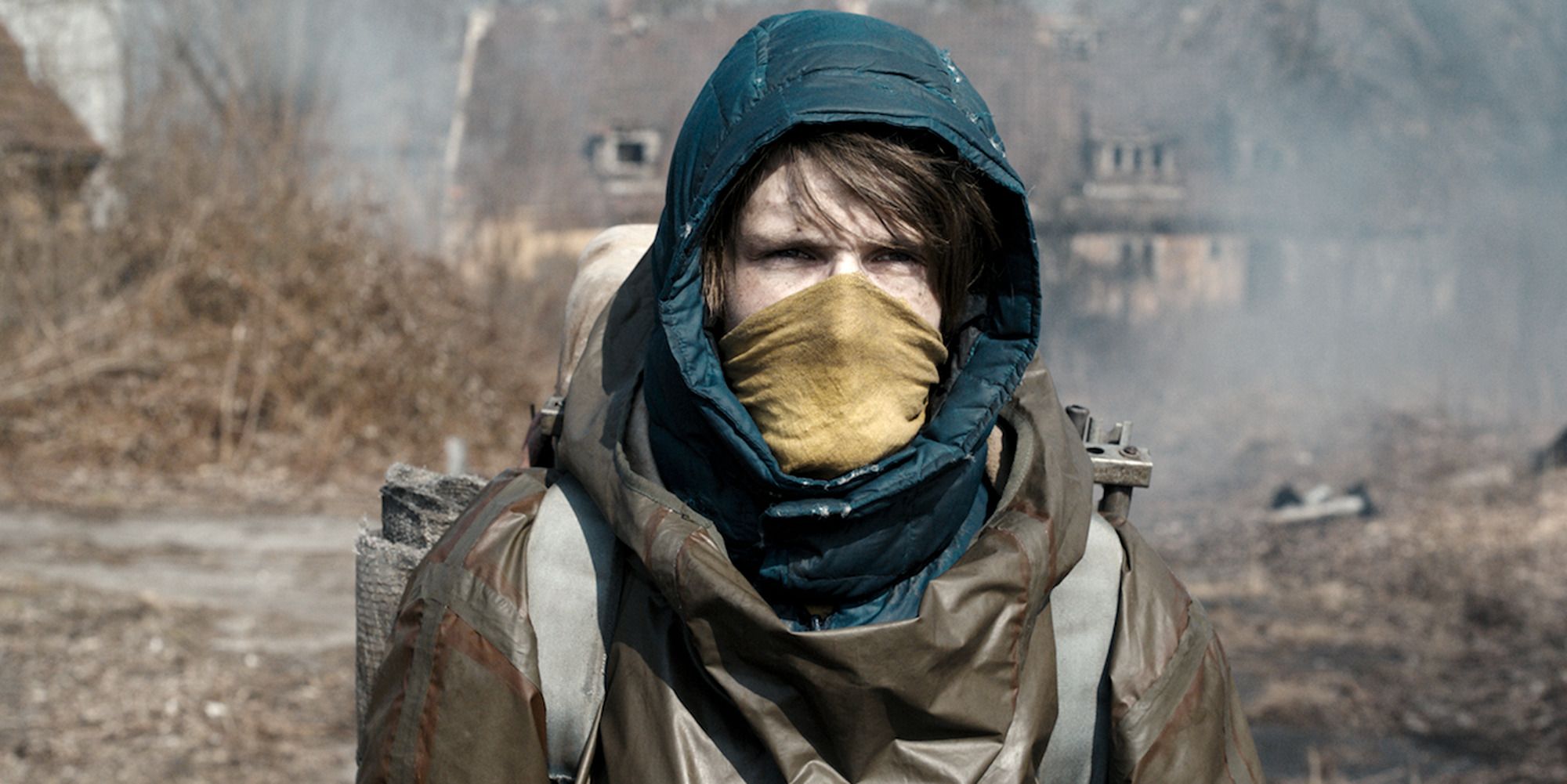 Hooded man wearing a yellow cloth mask, a barren wasteland behind him
