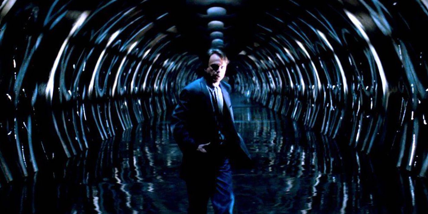 Sam Neill stands in a long, circular, mirrored hallway looking confused in In the Mouth of Madness