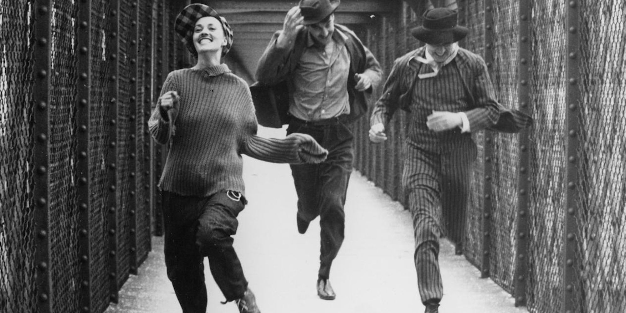 A smiling Catherine, Jules, and Jim run down a narrow path surrounded on either side by a wired fence. 