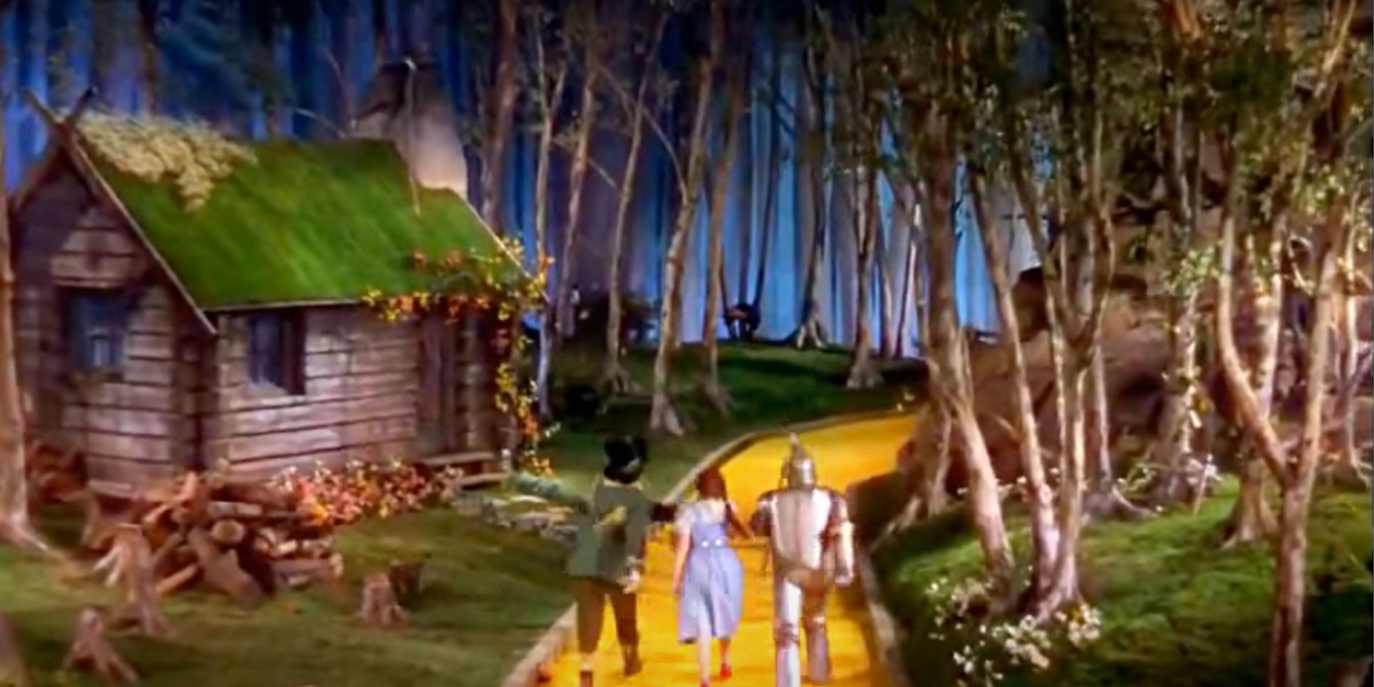 The Scarecrow, Dorothy, and the Tin Man skip merrily along the yellow brick road against a forest backdrop, with something moving in the background