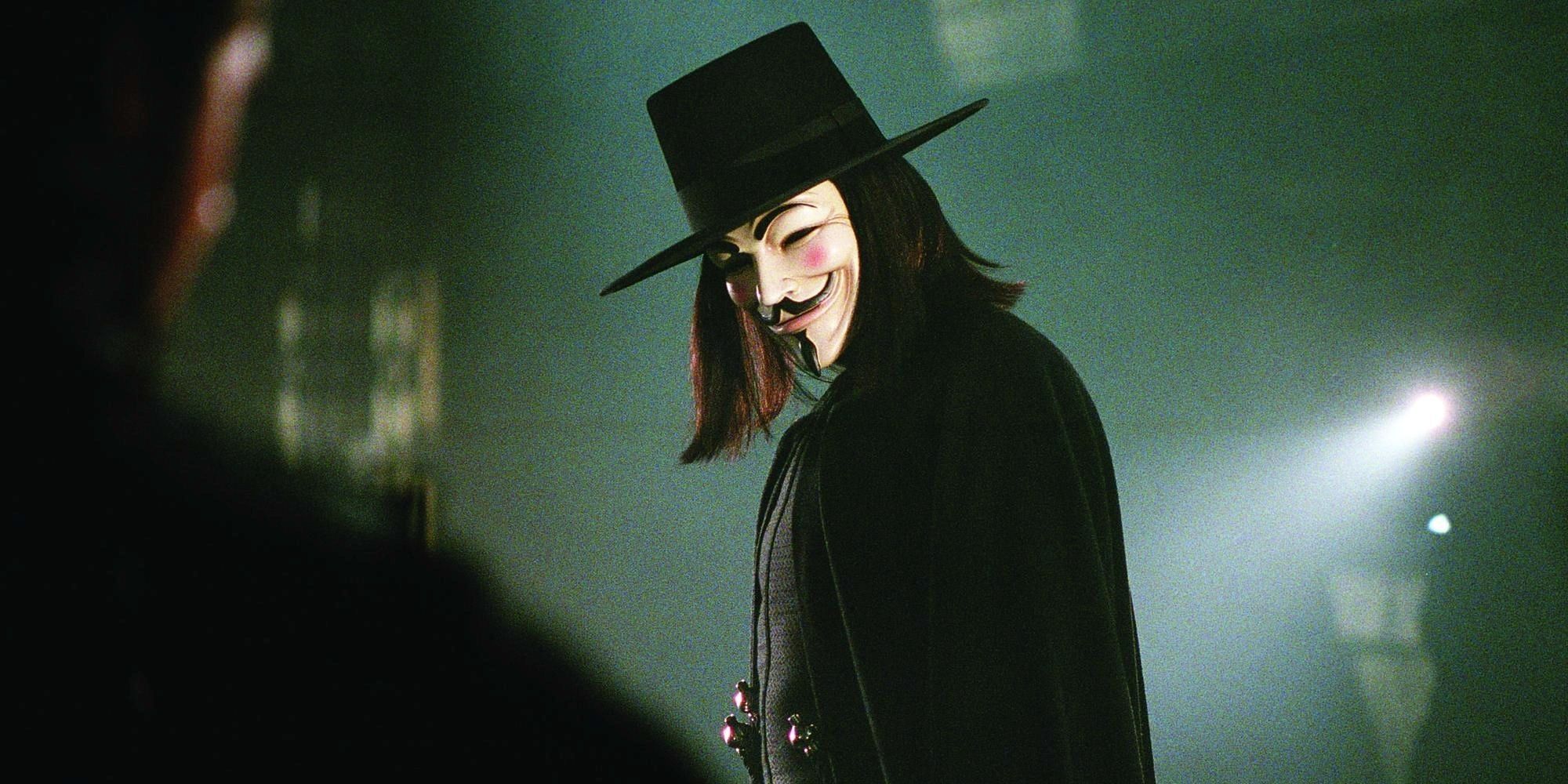 A man wearing a Guy Fawkes mask, a black hat, and a black cape stands surrounded by armed me.