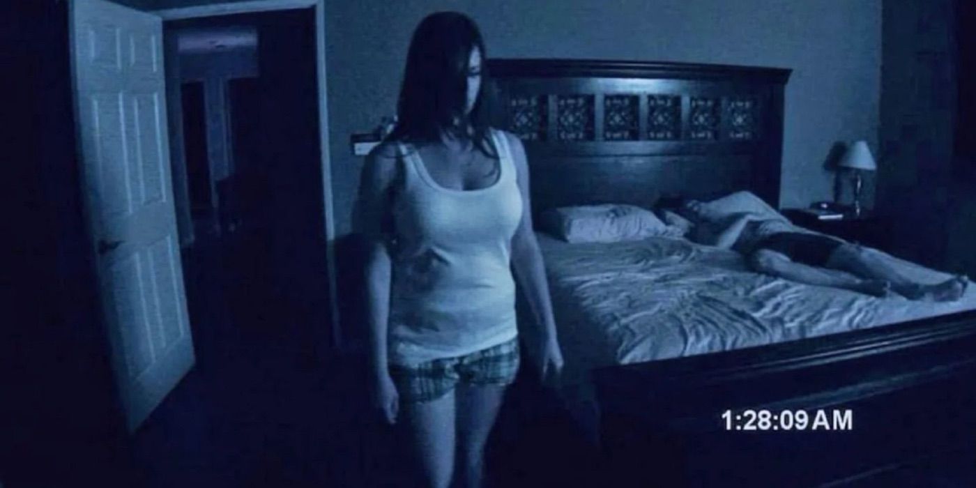 Une femme se tient debout et regarde attentivement pendant qu'un homme dort sur le lit derrière elle dans Paranormal Activity (2007).