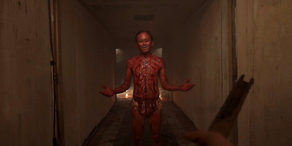 Still from VHS 2: A man stands smiling in a hallway, covered in blood with carvings on his chest.