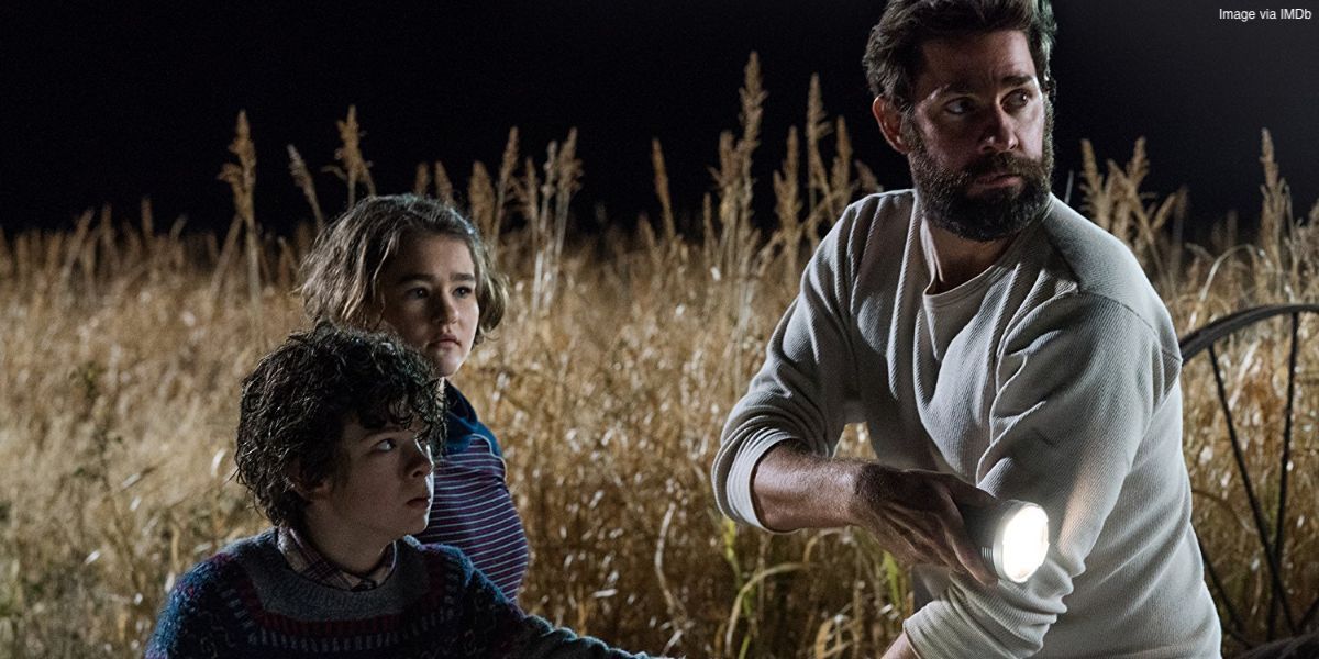 A man and two children in a wheat field looking in the same direction in the 2018 movie A Quiet Place