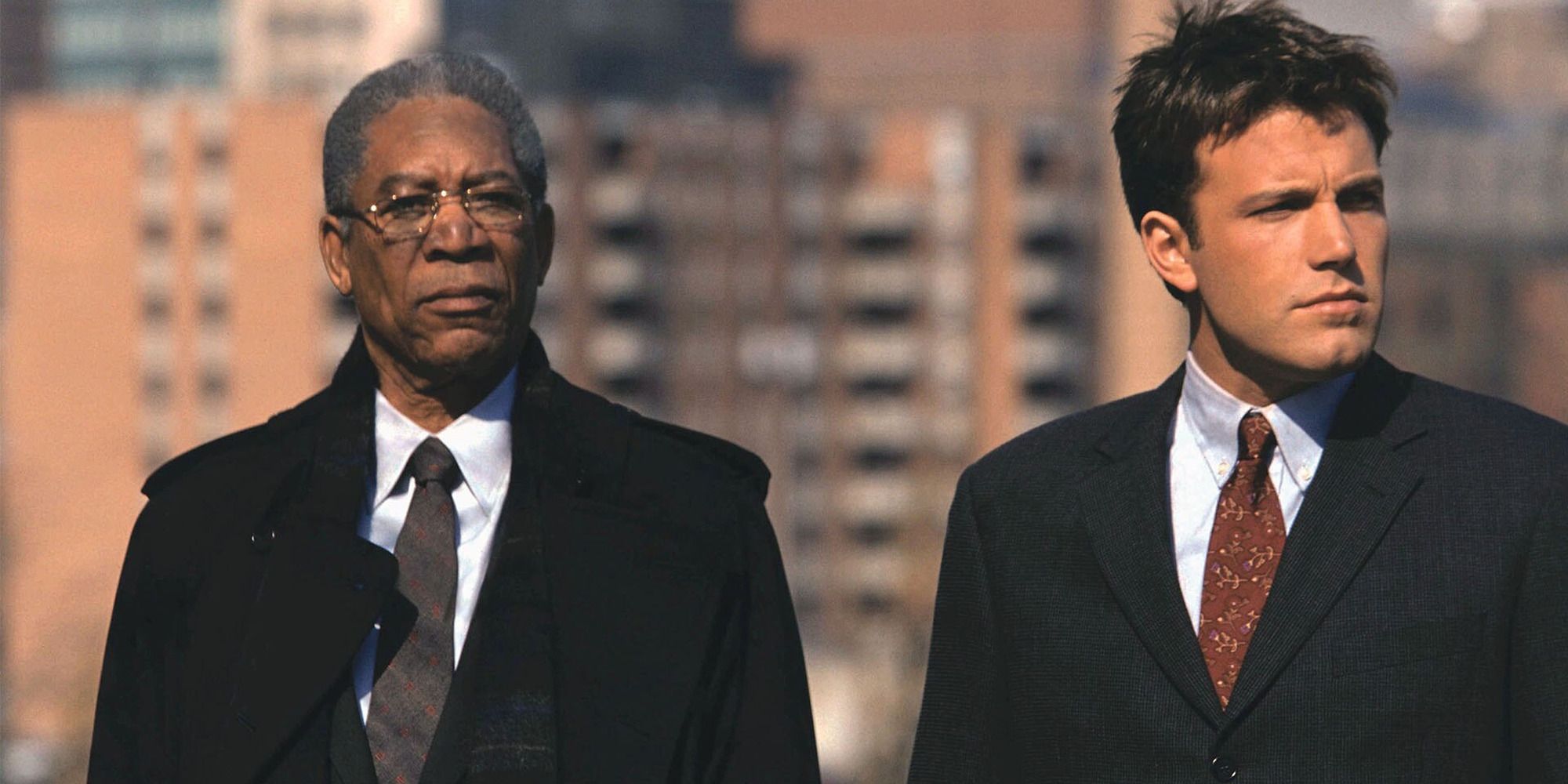 two men in suits standing atop a building with the city in the background