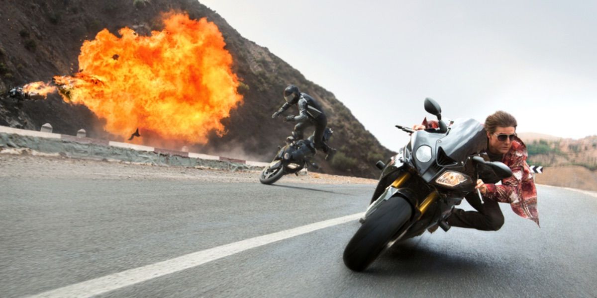 Ethan Hunt (Tom Cruise) races around a street corner on his motorcycle while pursuing enemies crash in the background.