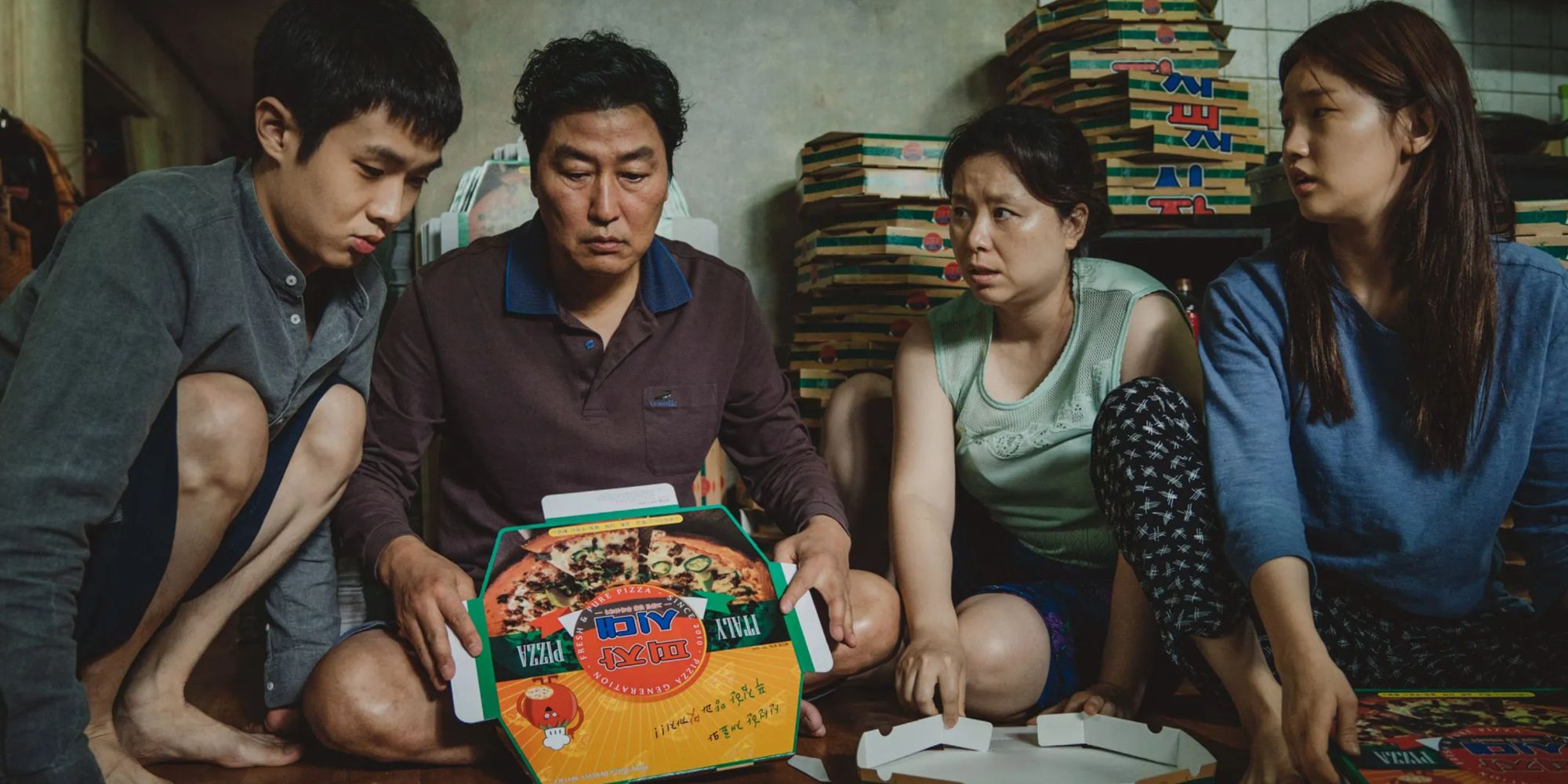 a family sits on the floor and looks at a cardboard box of pizza in Parasite