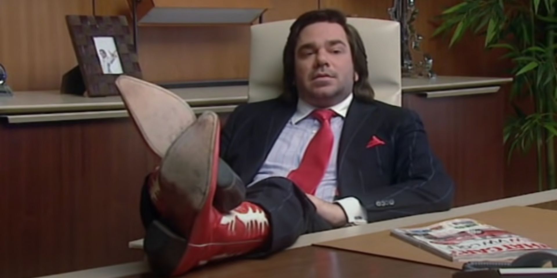 Douglas Reynholm (Matt Berry) sits back with his cowboy boots on the desk in The IT Crowd.