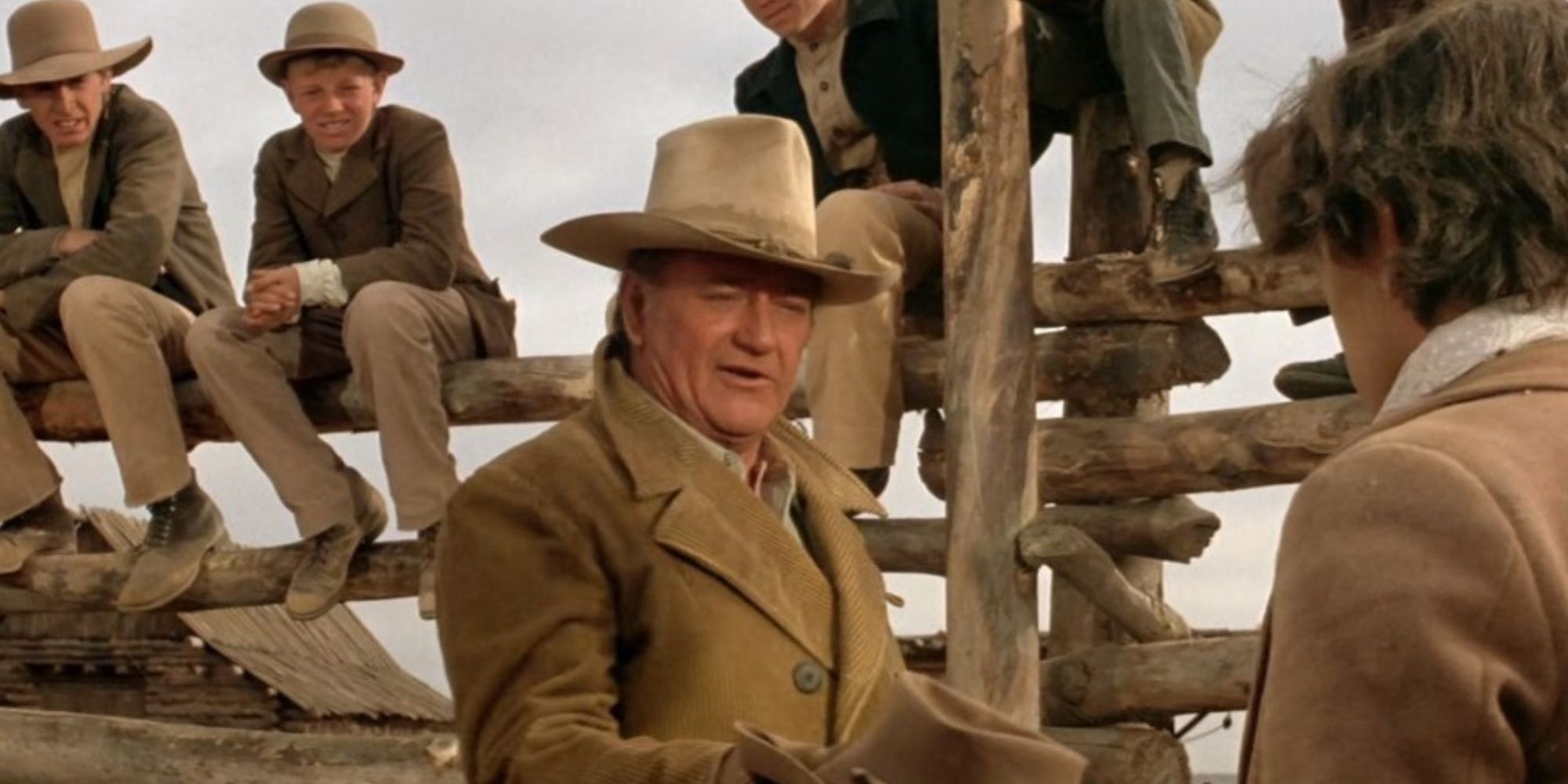 Boys sitting on a wooden fence behind Wil Andersen (John Wayne) who is speaking to Cimarron (A Martinez) in 'The Cowboys.'