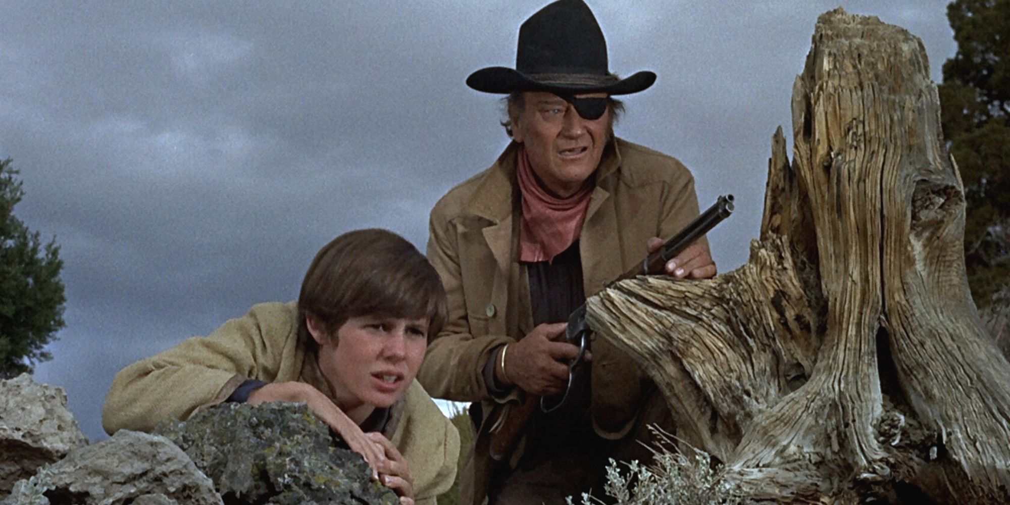 Kim Darby e John Wayne com uma espingarda agachados atrás das pedras olhando para algo em True Grit