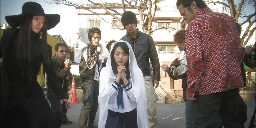 Une femme en train de prier, entourée de plusieurs personnes prêtes à la combattre dans 'Love Exposure'.