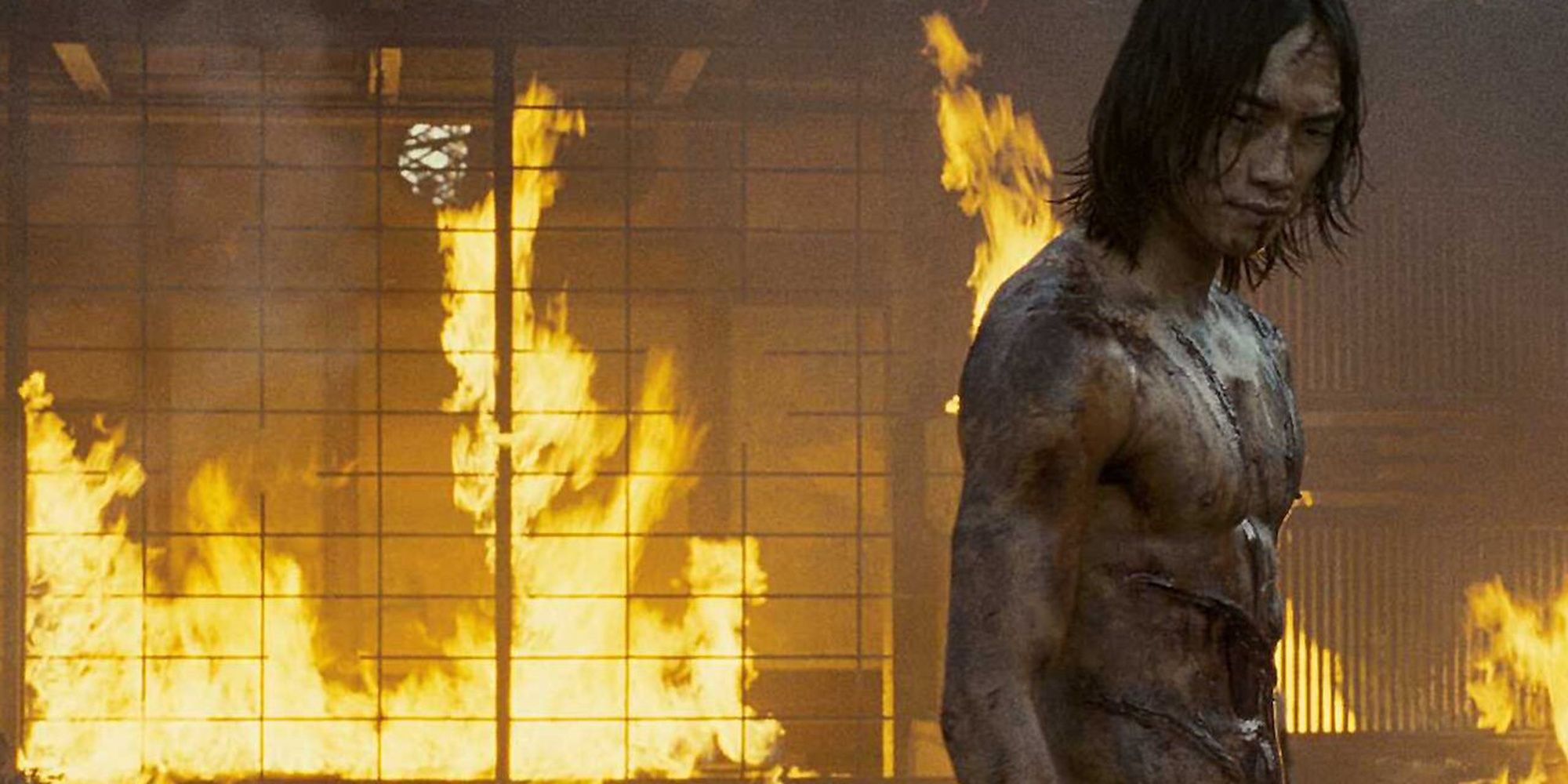 A shirtless and bloodied man standing in front of a burning building