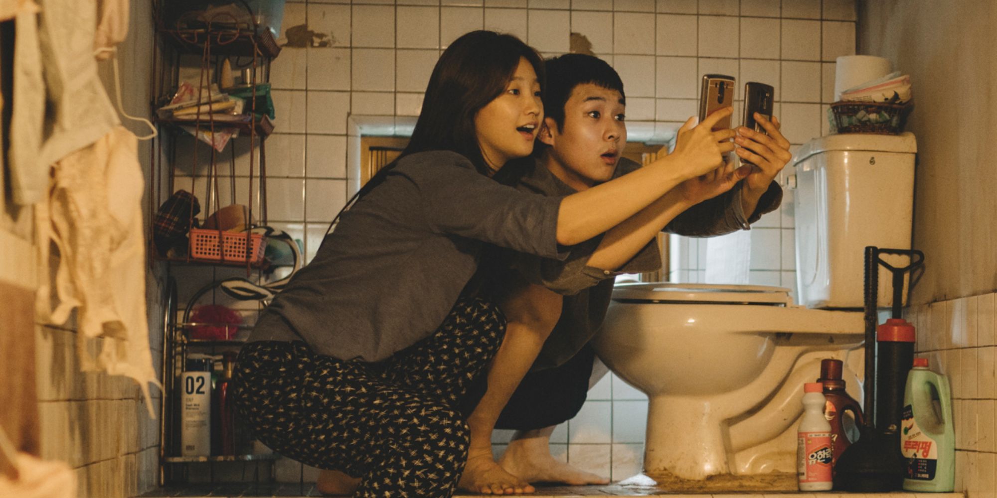 Choi Woo-shik and Park So-dam as Kim Ki-woo and Kim Ki-jung, holding their phones up by a toilet, trying to get cell service in Parasite