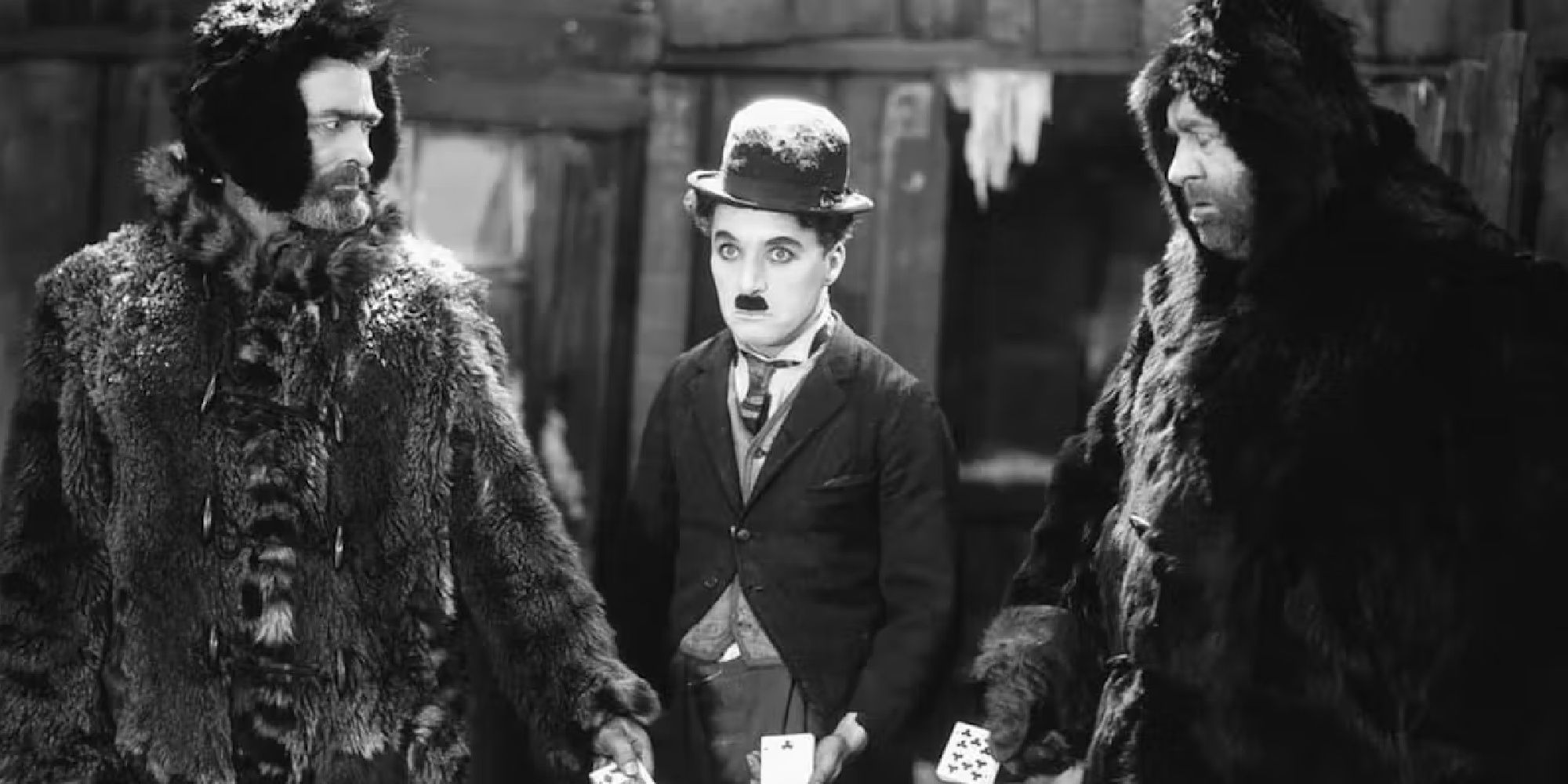 Charlie Chaplin estrelando 'The Gold Rush', dois homens vestindo casacos de pele ao lado dele.