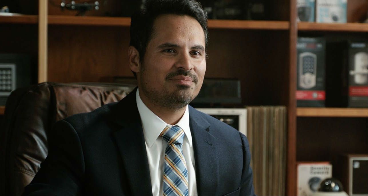 Michael Peña como Luis está sentado no escritório vestindo terno e gravata em 'Homem-Formiga e a Vespa'.