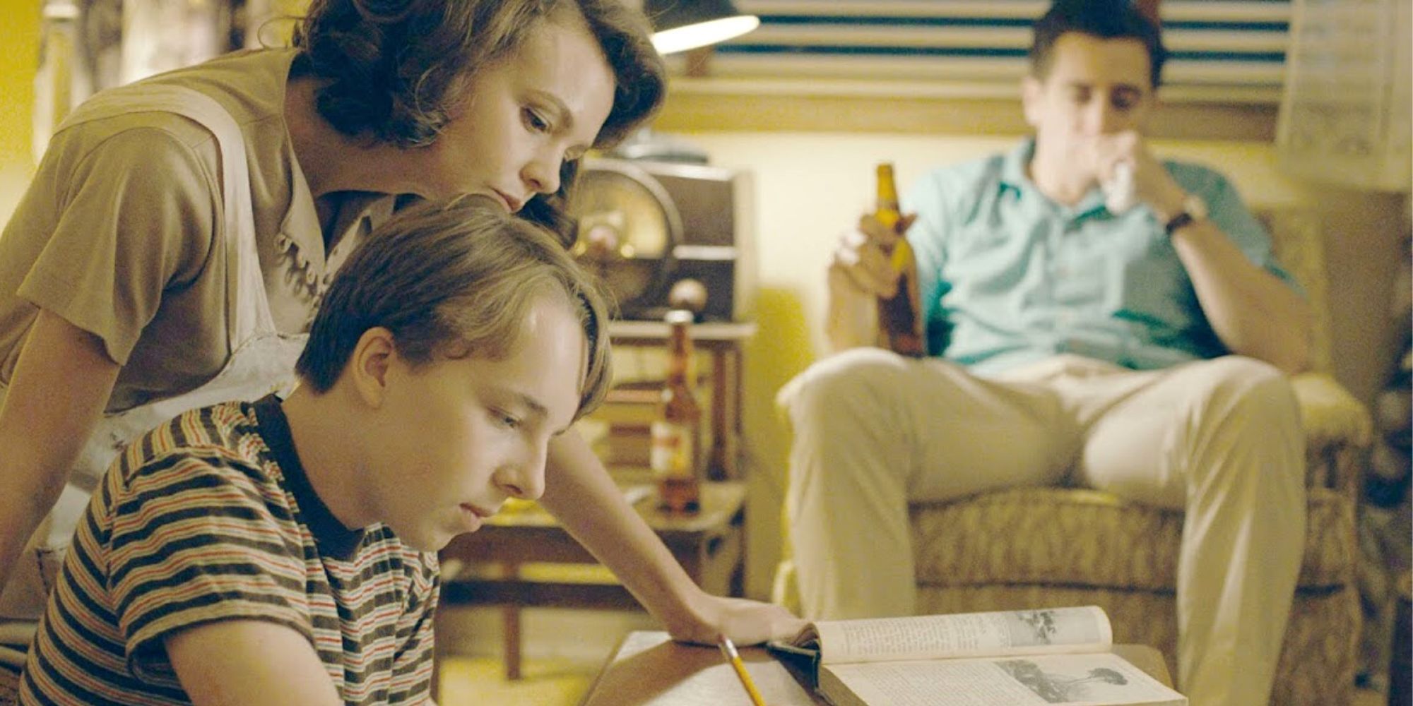 Carey Mulligan sitting next to Ed Oxenbould helping him with homework as Jake Gyllenhaal sits in the background in an armchair in Wildlife