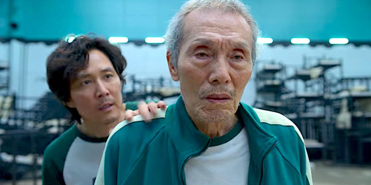 close up of Oh Il-Nam in Squid Game looking distressed, with Gi-Hun standing behind Oh Il-Nam and placing a hand on his shoulder looking concerned