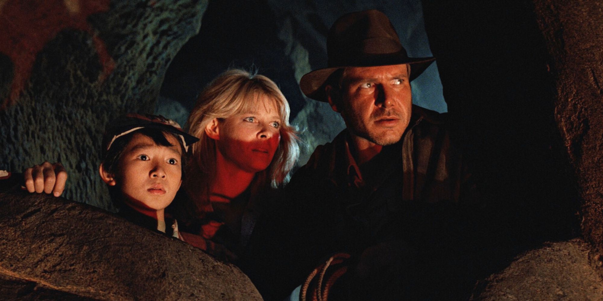 Ke Huy Quan, Kate Capshaw dan Harrison Ford berjongkok di balik bebatuan di sebuah gua yang sedikit mendongak ke arah Indiana Jones dan Temple of Doom