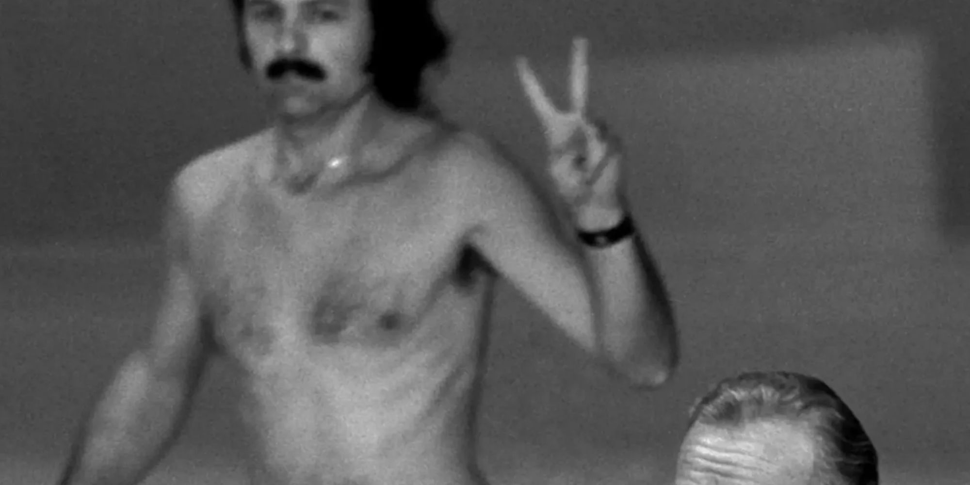 Shirtless man flashing peace sign; at bottom left of frame, forhead of man, rest of body is out of frame, 