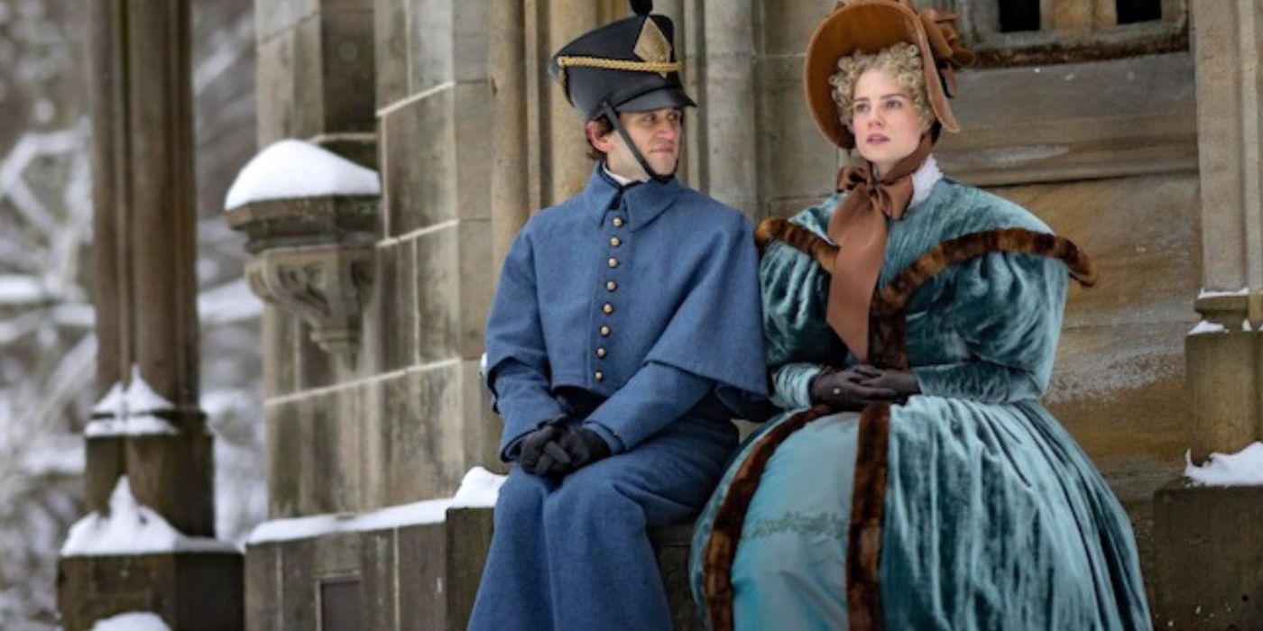 Harry Melling como Poe sentado ao lado de Lucy Boynton como Lea em um banco em frente a um mausoléu em The Pale Blue Eye