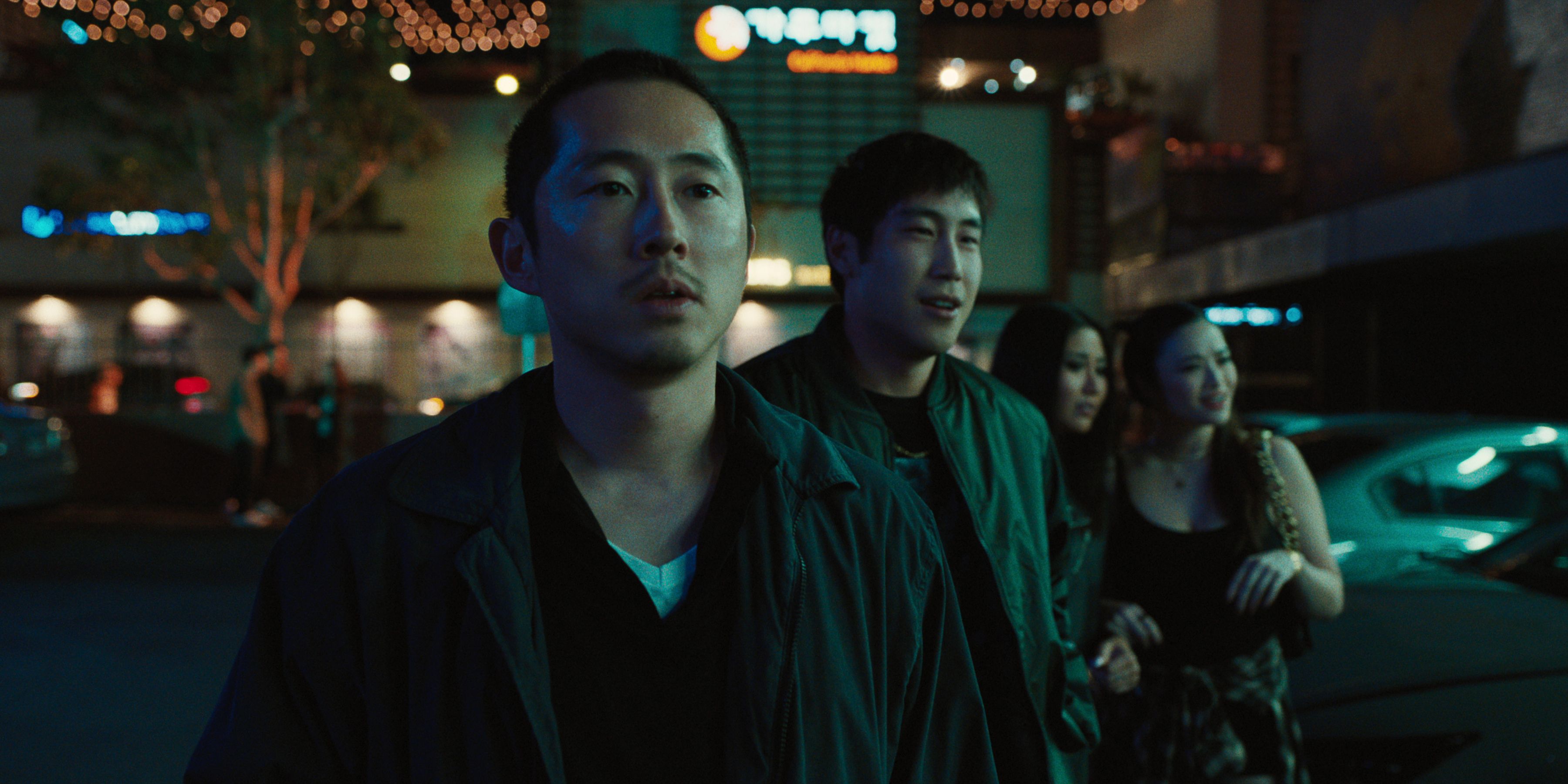 Steven Yeun (Danny Cho), debout dehors la nuit avec un groupe d'amis, regarde quelque chose au loin dans BEEF.