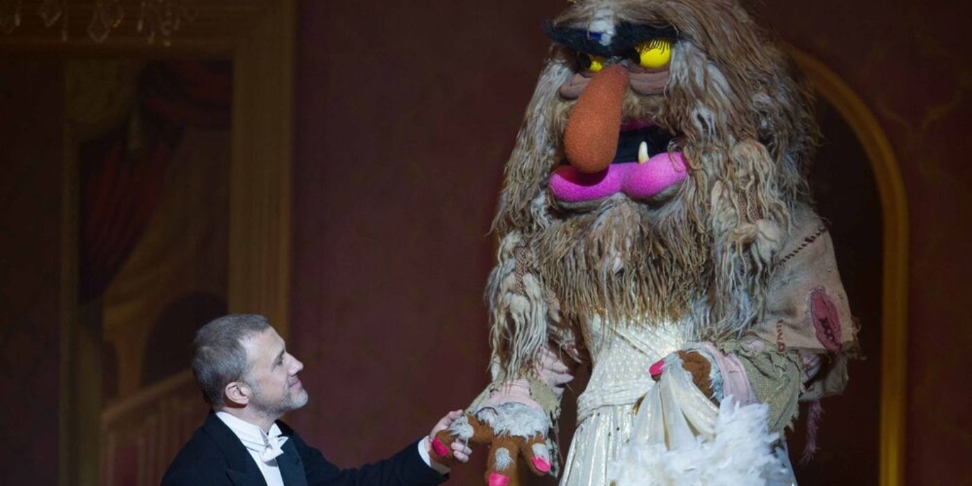 Christoph Waltz helping a large muppet wearing a wedding dress climb down a set of stairs in Muppets Most Wanted.