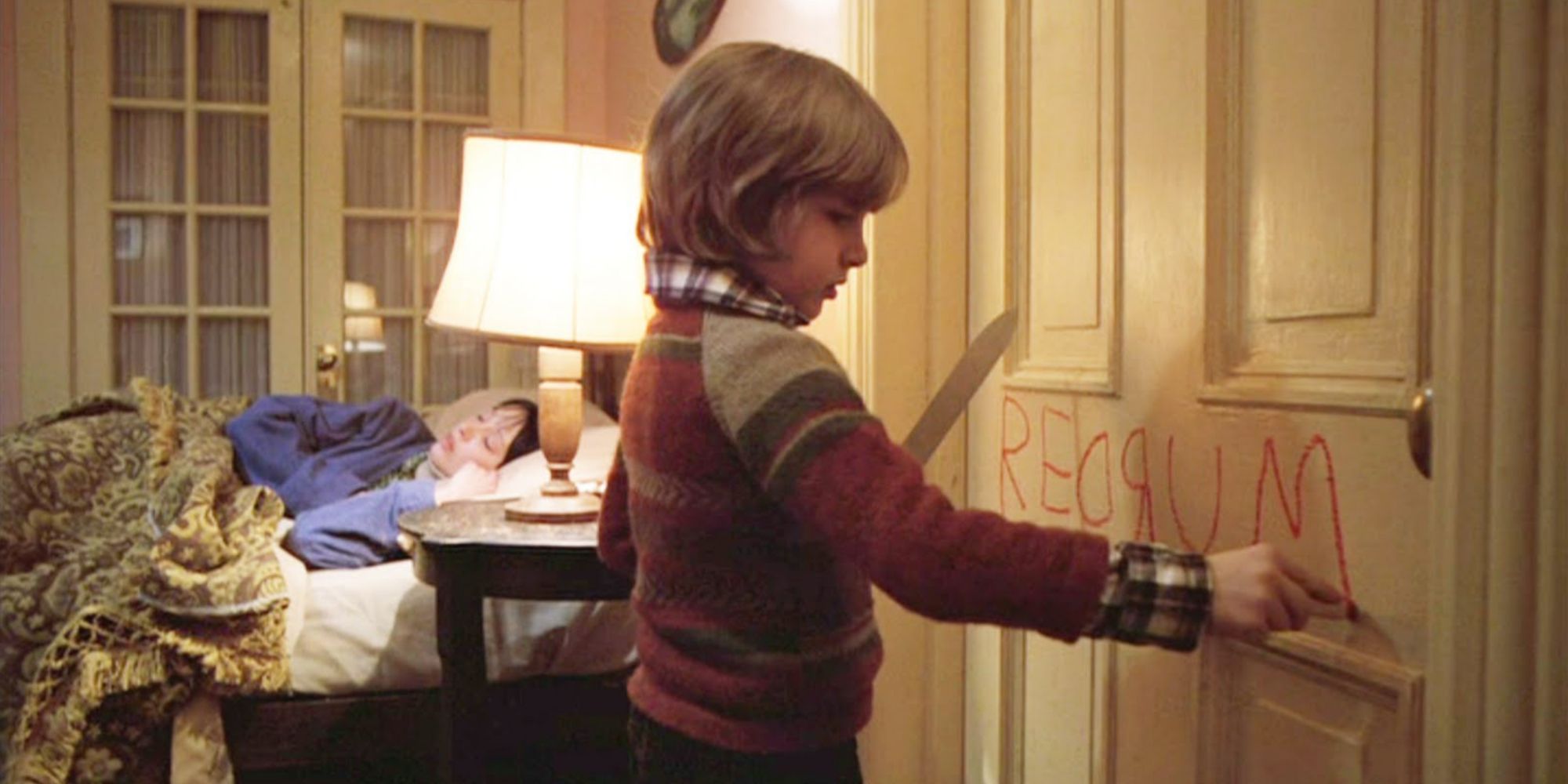 boy writing redrum on door holding knife: mother sleeping in bed in background