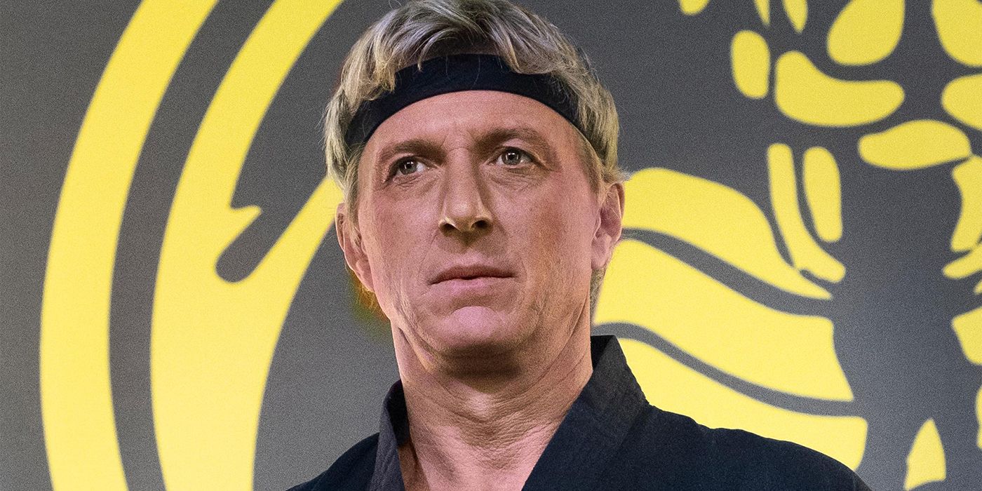 Johnny Lawrence in his dojo on Cobra Kai wearing a black headband, the Cobra Kai logo visible in part in the background.