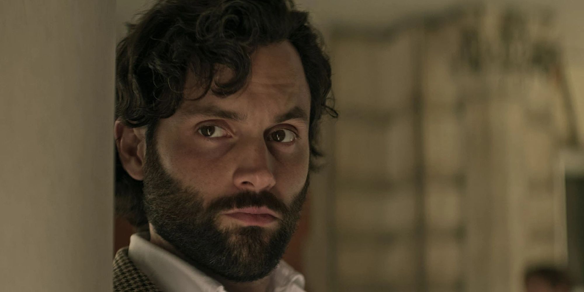 Penn Badgley as Joe Goldberg in 'You' standing at a doorway sneaking a peek at someone.