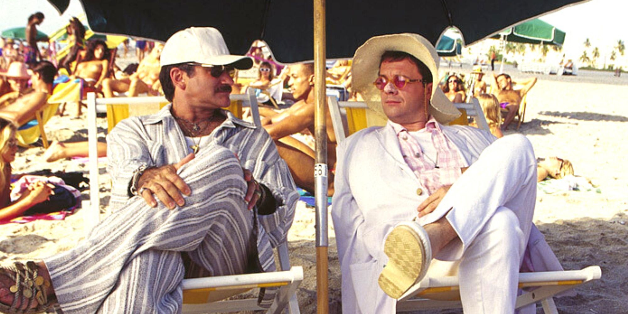 Robin Williams sentado junto a Nathan Lane en la playa en una jaula de pájaros