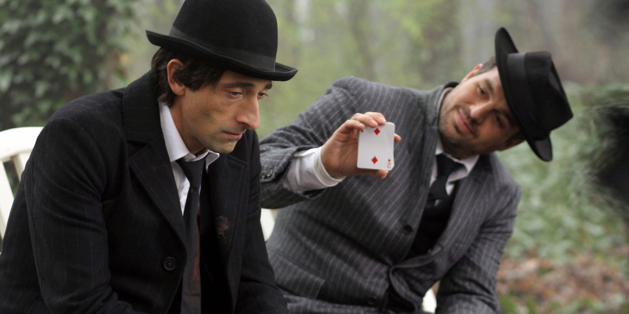 Two men sit on a park bench doing card tricks wearing top hats and suits in a scene from Brothers Bloom.