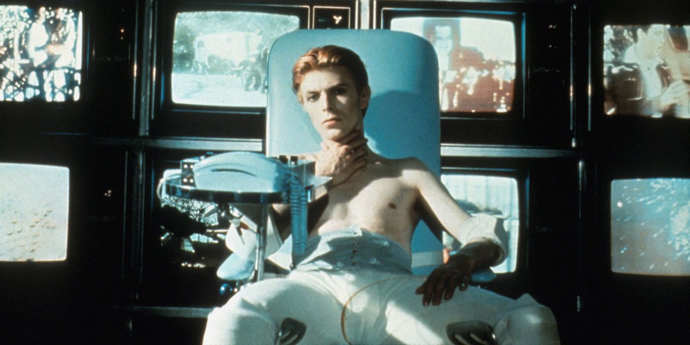 David Bowie sitting in a chair in front of a bunch of TVs, holding his throat, in The Man Who Fell to Earth