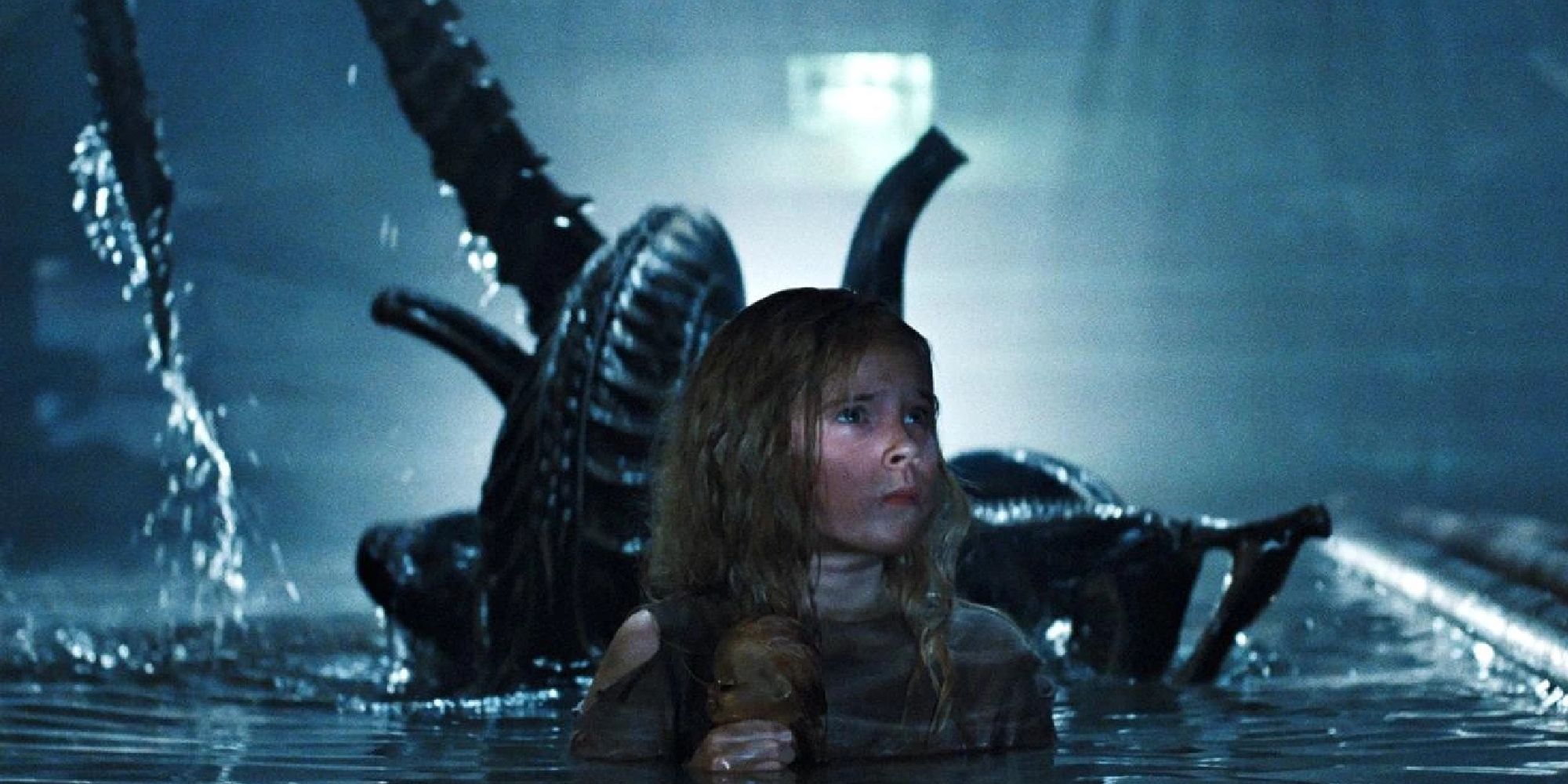 Newt (Carrie Henn) holding a baby doll whilst an alien stands behind her in 'Aliens'
