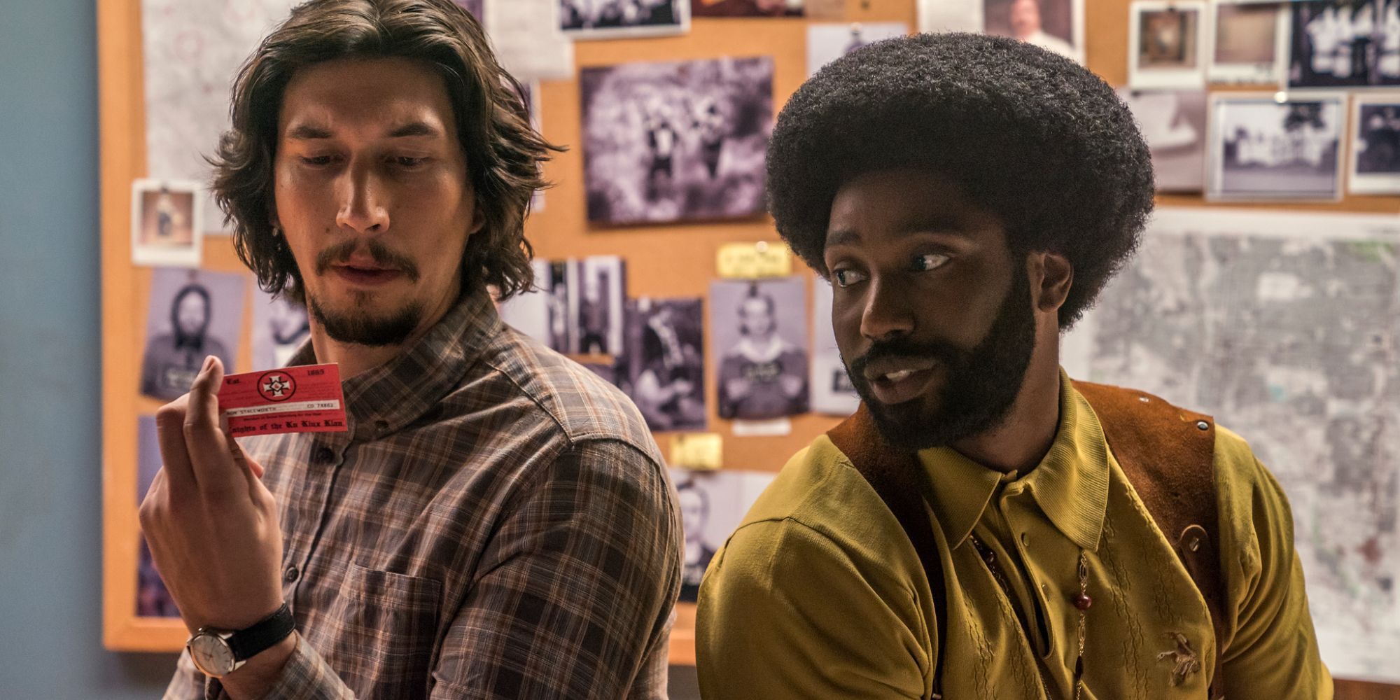 Adam Driver holding a KKK card sitting next to John David Washington in BlackKklansman.