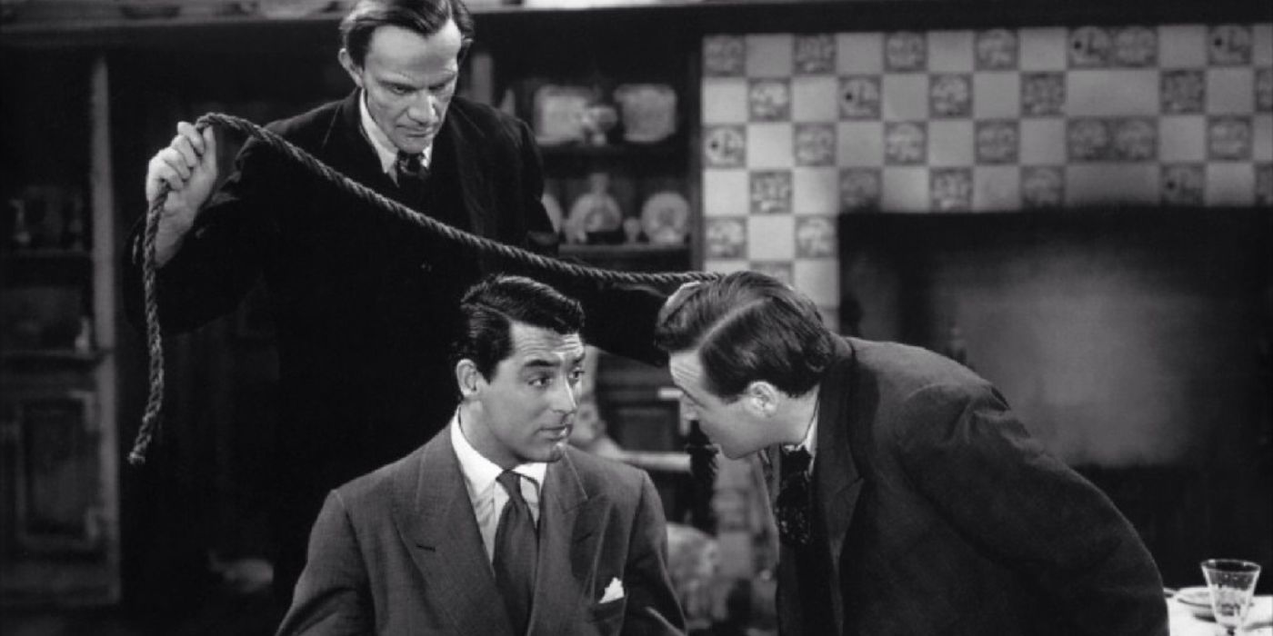 Raymond Massey debout derrière Cary Grant qui est assis sur une chaise et regarde Peter Lorre dans Arsenic et vieilles dentelles.