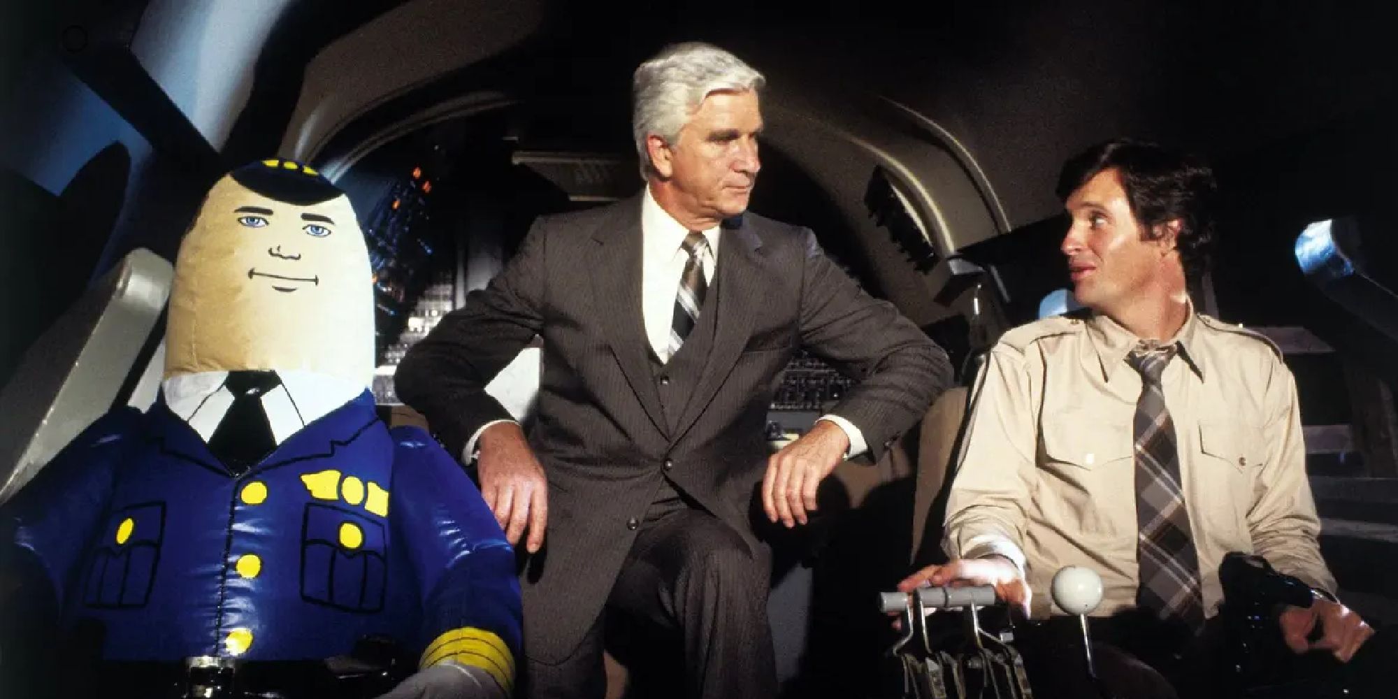 A doctor stand in the cockpit of an airplane having a discussion with the pilot while an inflatable co-pilot sits next to him.