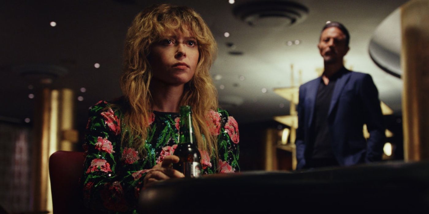 Natasha Lyonne wearing a floral dress holding a beer and looking at someone, Benjamin Bratt as Cliff in the background in a scene from Poker Face.