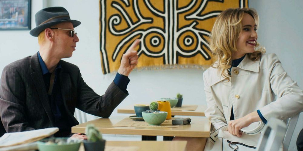 Une capture d'écran de Suki Waterhouse et Ewen Bremner assis à une table de café lors d'une interview dans Creation Stories (2021)
