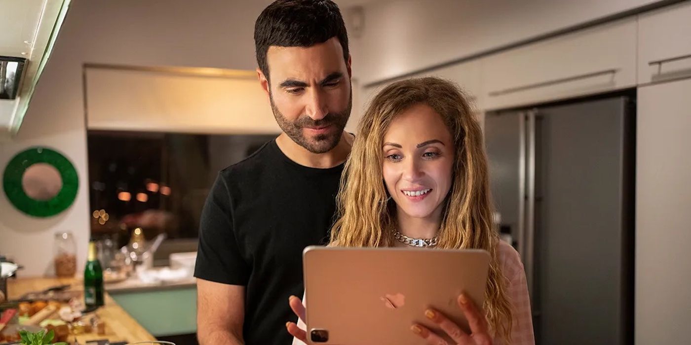 Roy, played by Brett Goldstein, looking over Keeley's, played by Juno Temple, shoulder as they look at pictures on her tablet in Ted Lasso Season 2