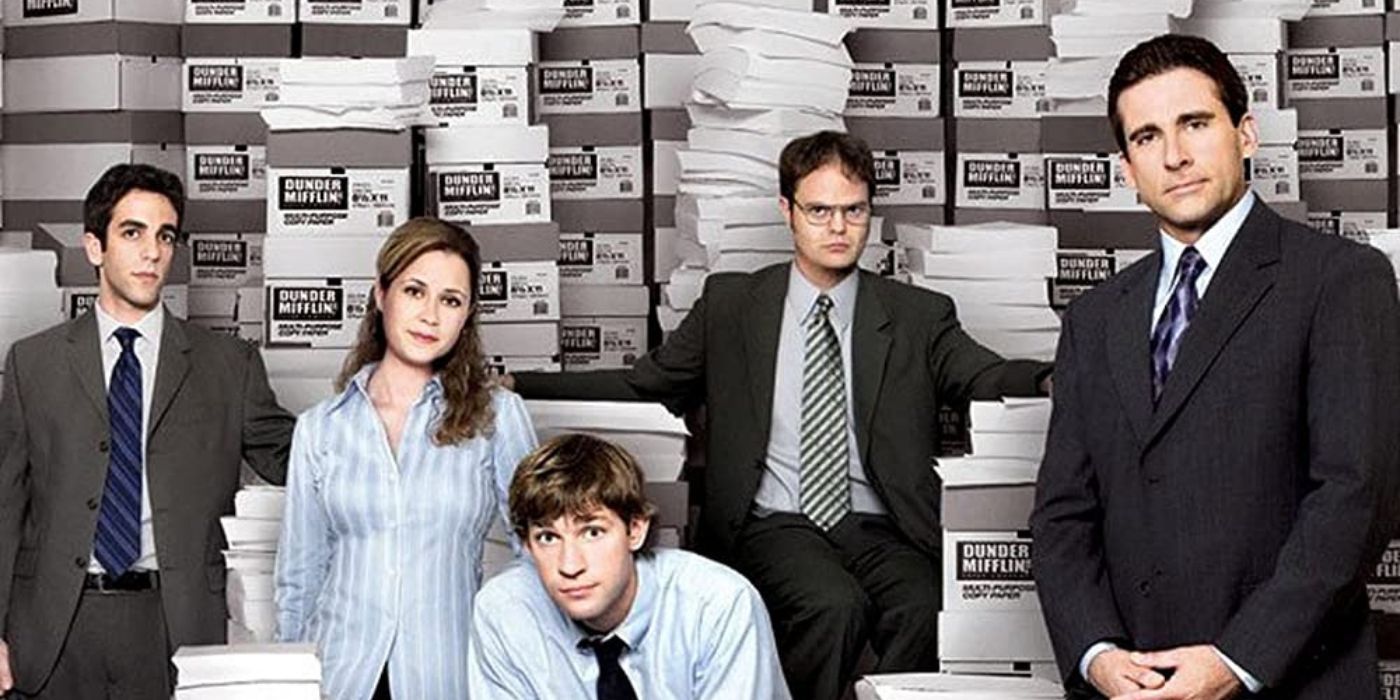 The main cast of The Office posing next to mountains of paper sheets.