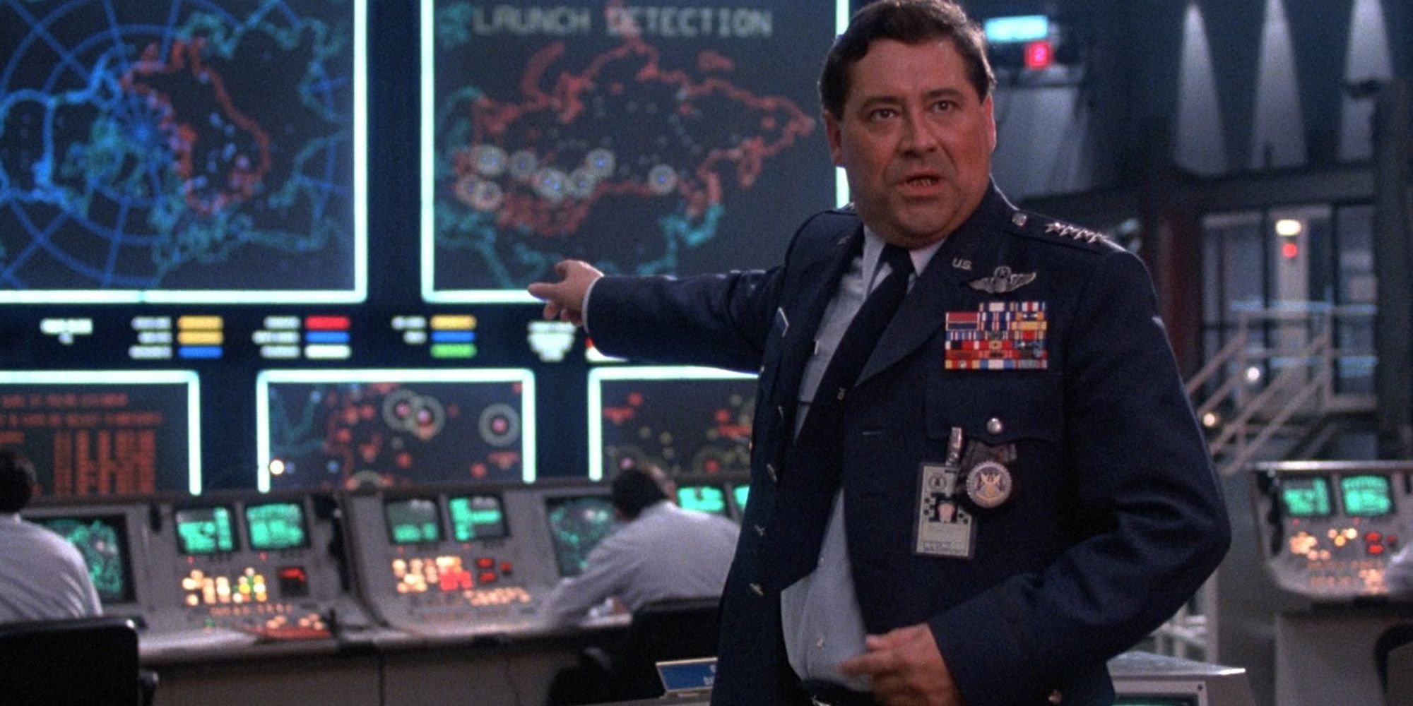 A military official points at several monitors while looking towards the camera in WarGames